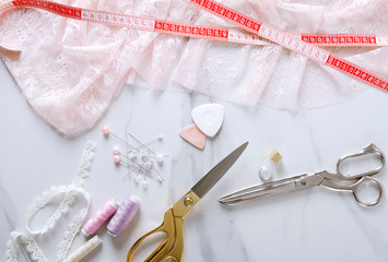 Top view of pink lace, measuring tape, golden and silver scissors, sewing pins on the white marble table