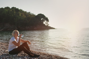 Mature woman drinking water during sunset after jogging. Senior lady sitting on the beach against...
