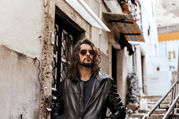 Young bearded and long haired man wearing leather jacket and sunglases walking on the southern european street
