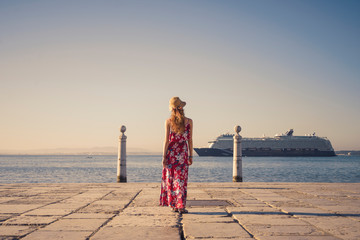 Young blonde female tourist wearing red generic sundress and a hat is looking at cruise ship going...