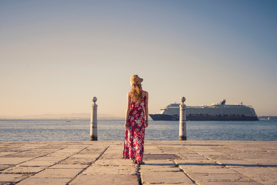 Young Blonde Female Tourist Wearing Red Generic Sundress And A Hat Is Looking At Cruise Ship Going To The Port In Lisbon From Atlantic Ocean, Portugal