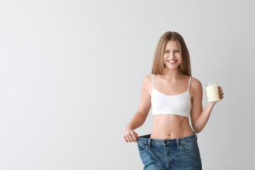 Beautiful young woman in loose jeans and with jar of coconut oil on light background. Weight loss...
