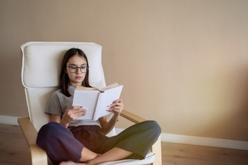 Cute teenage girl sitting on sofa at home and reading book. Education, children and school concept,...