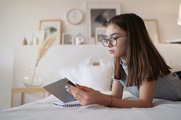 Cute teenager girl using tablet lying on bed at home. Young pretty girl wearing glasses studying online, distance learning, self education
