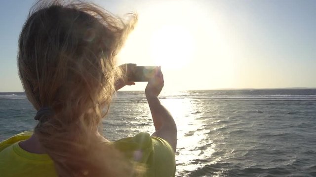 Woman shooting video of amazing sunset over windy blue sea water using her modern mobile smartphone. Back view slow motion of blond wavy female hair moving in strong wind. Egypt, Red sea.