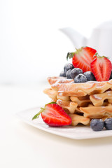 Stack of delicious . waffles with blueberries and sliced strawberries .close-up on white table with white.teapot from the back, copy space.