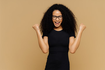 Studio shot of overjoyed cheering woman with curly hair, raises clenched fists, celebrates success...