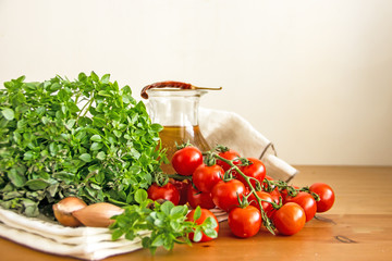 Cherry tomatoes on a sprig next to the greens of fresh basil, shallot onions, chili pepper, olive oil in a transparent decanter on a white napkin on a wooden table. Copy space.