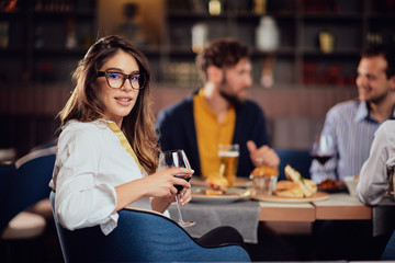 Portrait of gorgeous brunette looking at camera and holding glass with red wine while sitting at restaurat. In background friends eating diner.