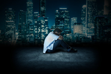 Close up shot of a young man sitting and crying on a floor of the roof top of a corporate building. Side view shot of a student very depressed. Sad teenage boy sitting on ground thinking at night.