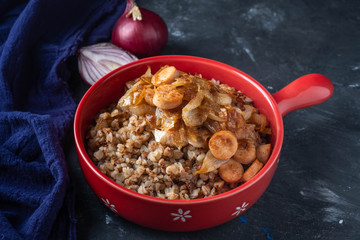 buckwheat porridge with sausages and fried onions in a red pan on a dark background, copy space, Close-up
