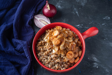 buckwheat porridge with sausages and fried onions in a red pan on a dark background, copy space, Close-up