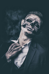 Stylish and beautiful, emotional young man with skeleton makeup in a strict suit against the background of smoke and dark background to Halloween