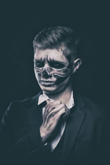 Stylish and beautiful, emotional young man with skeleton makeup in a strict suit against the background of smoke and dark background to Halloween