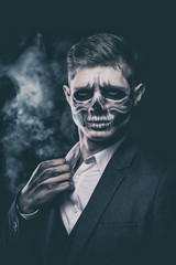 Stylish and beautiful, emotional young man with skeleton makeup in a strict suit against the background of smoke and dark background to Halloween