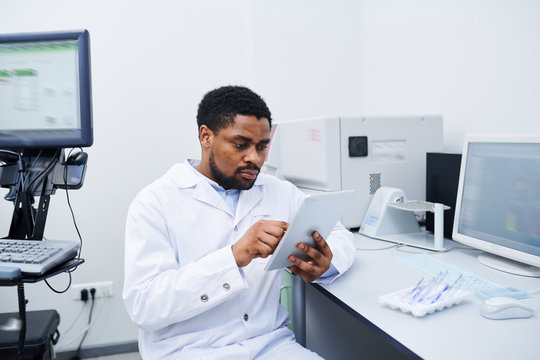 Serious Concentrated Young Black Medical Scientist With Beard Sitting At Desk And Using Digital Tablet While Analyzing Data In Laboratory