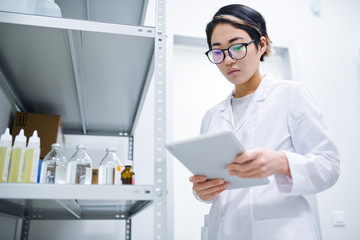 Serious concentrated young Asian lady in white coat wearing eyeglasses using digital tablet while searching for information about medication in database, she working in medical storage room