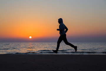 Alone man running on sand beach. Beautiful colorful sunset sky. Sportsman in tracksuit with hood. Enjoying sport in evening. Outdoor workout. Daily active lifestyle. Side view. 