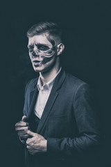 Stylish and beautiful, emotional young man with skeleton makeup in a strict suit against the background of smoke and dark background to Halloween