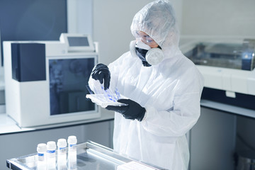Busy infectious disease scientist in biohazard suit and respiratory mask standing at table with...