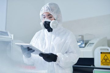 Busy laboratory biologist in protective costume and respiratory mask using digital tablet while...