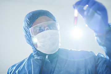 Concentrated black infectious disease scientist in biohazard suit and mask holding test tube and analyzing virus-infected blood sample