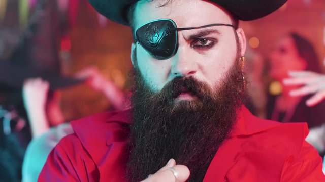Portrait of a young man dressed in pirate costume with an evil look and big black beard. People dancing in the background at a halloween party.
