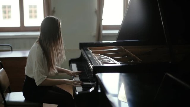 Beautiful young woman playing the black piano on the background of the window in slow motion. Pianist's face covers her long hair