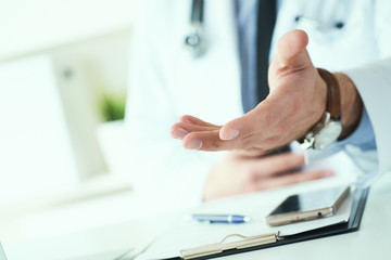 Male doctor making welcome gesture, politely inviting patient to sit down in medical office. Photo with depth of field.