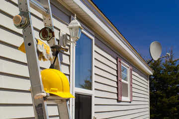 Yellow hard hat, work gloves and hammer on ladder with house and wireless dish background.