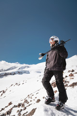 Fototapeta na wymiar Growth Portrait bearded male skier aged against background of snow-capped Caucasus mountains. An adult man wearing ski googles mask and helmet skis on his shoulder looks mountains. Ski resort concept