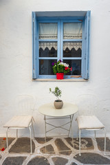 Fototapeta na wymiar White house facade with blue window, table and chairs in Naoussa town, Paros island, Cyclades