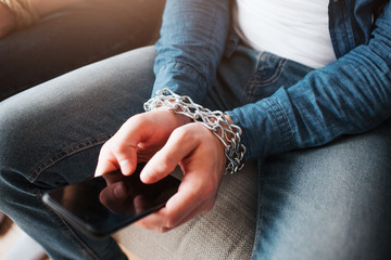 Young man have social media addiction. Phone in hands. Chains around wrist. Hostage of social media. Sitting on sofa. Cut view.