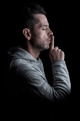 Side studio portrait of a man with closed eyes, with finger on lips making silence gesture. Isolated on black background. Vertical.