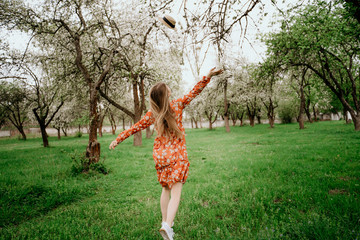 Young beautiful blonde woman in blooming garden. Spring trees in bloom. Orange dress and straw hat.