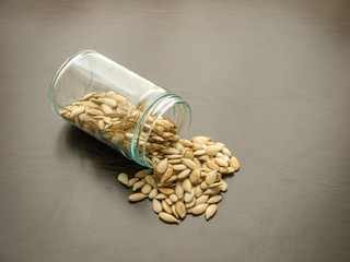 Unpeeled pumpkin seeds are poured from a glass jar on brown wooden surface