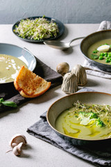 Variety of green vegetable soup asparagus, broccoli and pea, decorated by greens, vegetables, quail eggs, cream, olive oil, in ceramic bowls over grey spotted table with ingredients above.