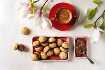 Baci di dama homemade italian hazelnut biscuits cookies with chocolate cream served in ceramic plate with red cup of espresso coffee and magnolia flowers over white marble background. Flat lay, space