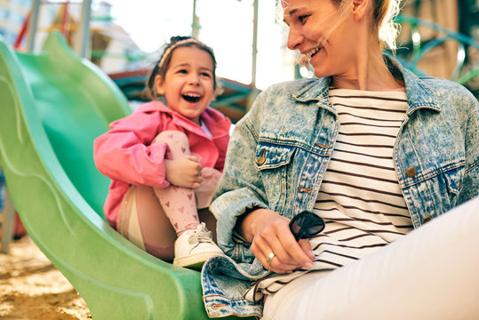 Outdoor image of happy cute little girl laughing with her beautiful mother spending time together at slide on the playground. Young woman and daughter feeling happy, loving each other outside.