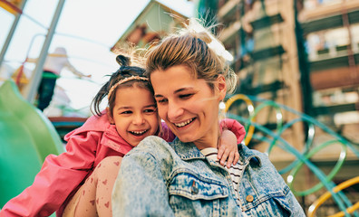 Obraz premium Outdoor portrait of happy cute little girl smiling and looking to her beautiful mother spending time together at playground. Young woman and daughter feeling happy, loving each other outside.