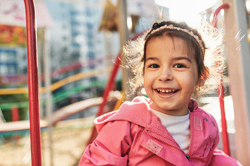 Outdoor closeup portrait of happy little girl smiling broadly, wearing pink outfit posing outside. Cheerful toddler kid playing at the playground in the park. Happy childhood and education concept.