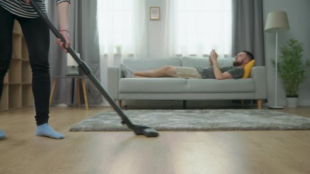Tired wife vacuuming parquet floor and her husband play on smartphone on sofa