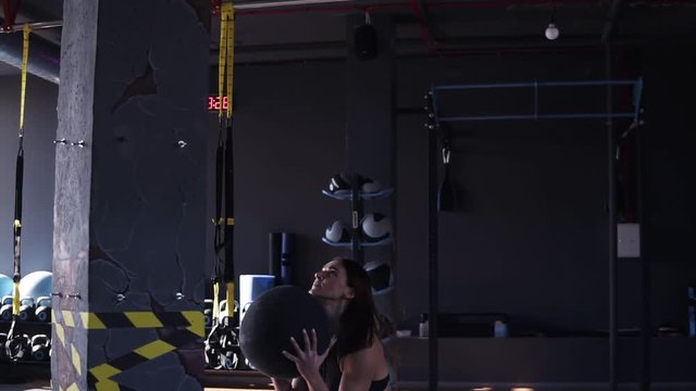 Sideview footage of a young fit woman in black leeggings and bra exercising by throwing a medicine ball up against a wall in a dark coloured modern gym full of equipment on the background