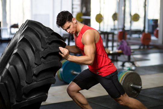 A sportsman in sportswear pushing immense wheel, isolated on a blurry gym background.