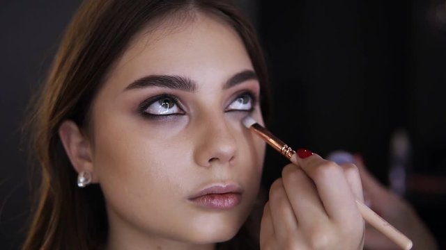 Portrait of young woman while makeup eyes in cosmetic studio. Makeup artist using brush to apply bright eyeshadow on low eyelid. Stylish makeup, beauty concept