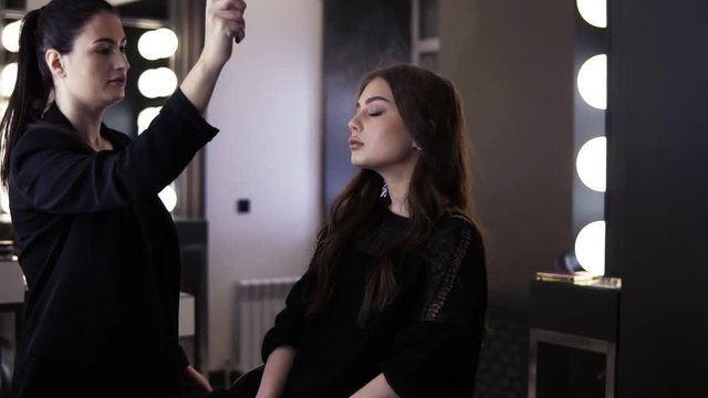 Makeup artist with black pony tail uses a make up finishing spray and gently sprays it on her client's face from a distance. Gorgeous young lady in black dress with finished make up and hairso