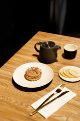 Gray teapot on a wooden table in a cafe. Black mug for tea. Dessert on a white plate and sliced lemon on a small saucer, golden cutlery a fork with a knife on a napkin. 