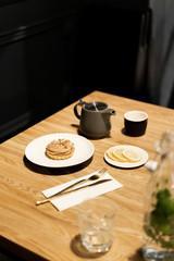 Gray teapot on a wooden table in a cafe. Black mug for tea. Dessert on a white plate and sliced lemon on a small saucer, golden cutlery a fork with a knife on a napkin. Glass for water and a carafe.