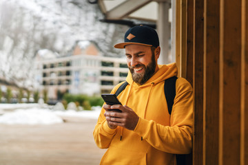 Front view. Bearded male tourist in yellow hoodie and cap with black backpack stands outdoors and using smartphone. Lifestyle.