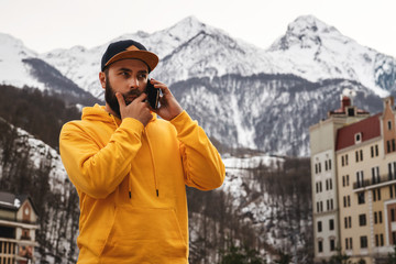 Bearded male tourist in yellow hoodie and cap stands on background of high snowy mountains, talking on mobile phone. Lifestyle.
